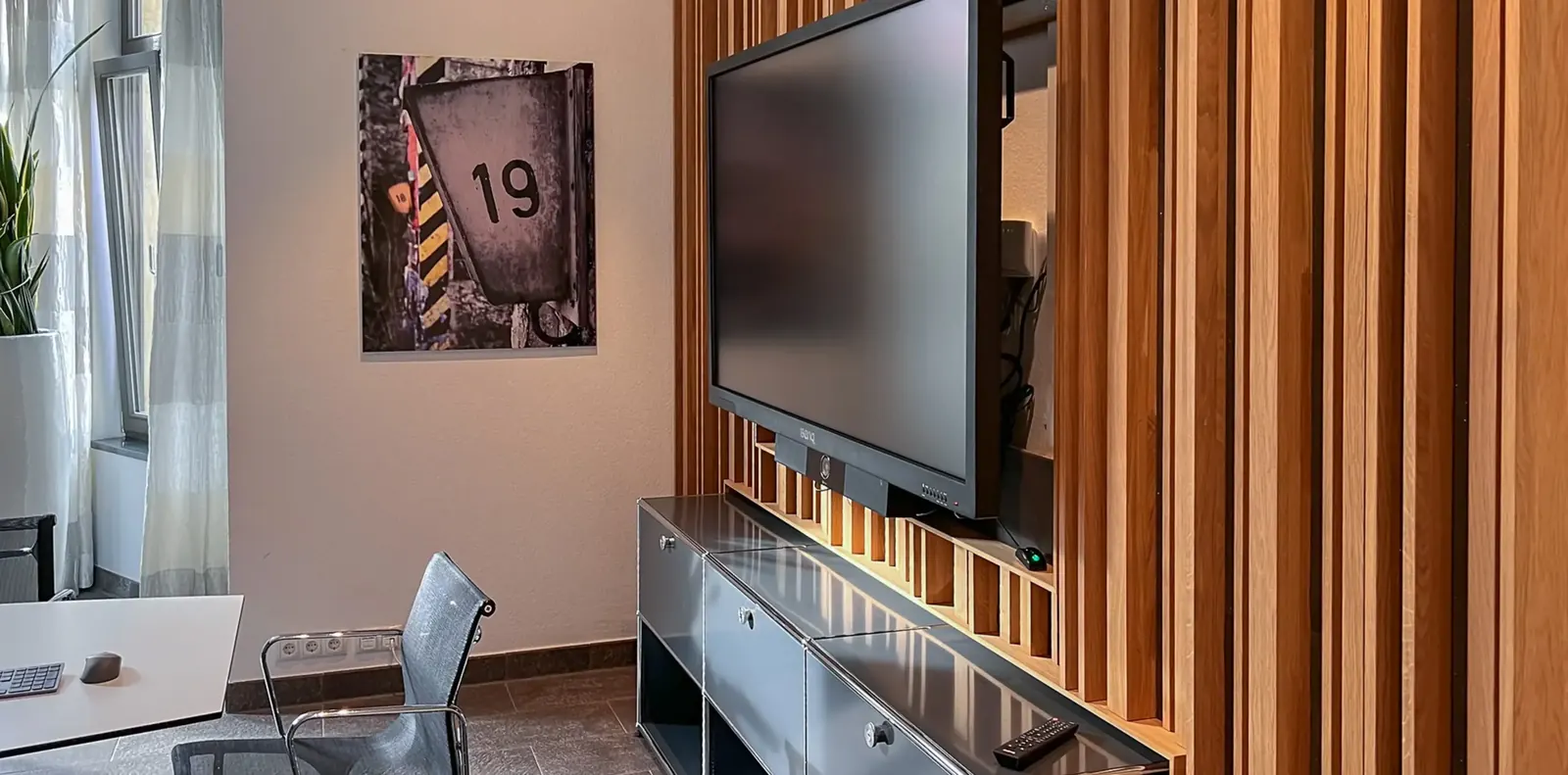 Besprechungsraum mit Holzlamellenwand, Bildschirm auf anthrazitfarbenem USM-Lowboard, weißem Tisch und Bürostuhl mit Netzbespannung.