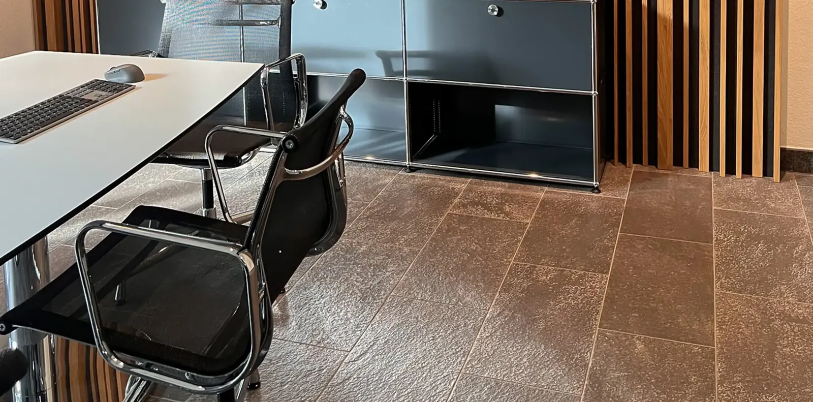 Weißer Arbeitstisch mit Tastatur und schwarzen Netzstühlen mit Chromarmlehnen auf Steinfliesen, dahinter USM-Regal und Holzlamellen.