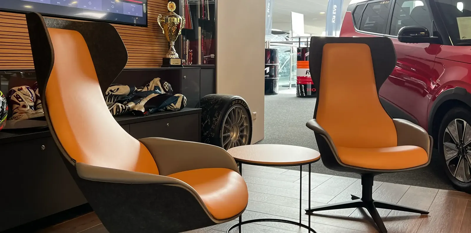 Orange-anthrazitfarbene Loungesessel mit Sternfuß und kleiner Beistelltisch im Autohaus, rechts ein roter Neuwagen.