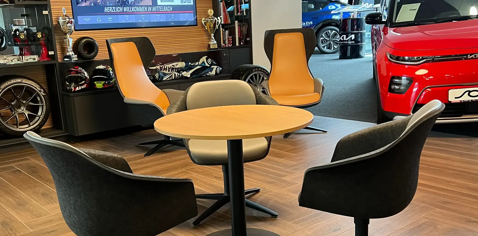 Runder Holztisch mit dunkelgrauen und beigen Schalensesseln, dahinter orangefarbene Loungesessel und ein roter Wagen.