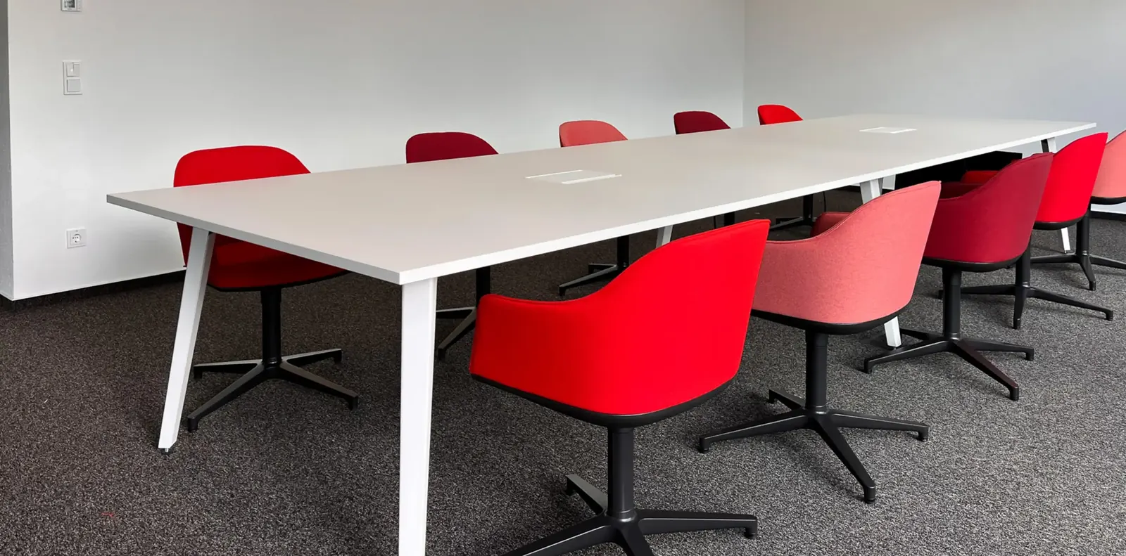 Konferenztisch mit weißen schrägen Beinen und Polsterstühlen in Rot, Rosa und Bordeaux auf grauem Teppichboden.