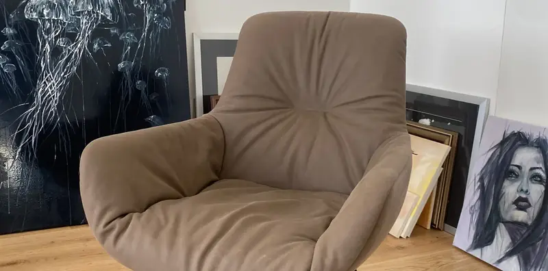 Taupefarbener Loungesessel mit gesteppter Polsterung vor an die Wand gelehnten Gemälden.