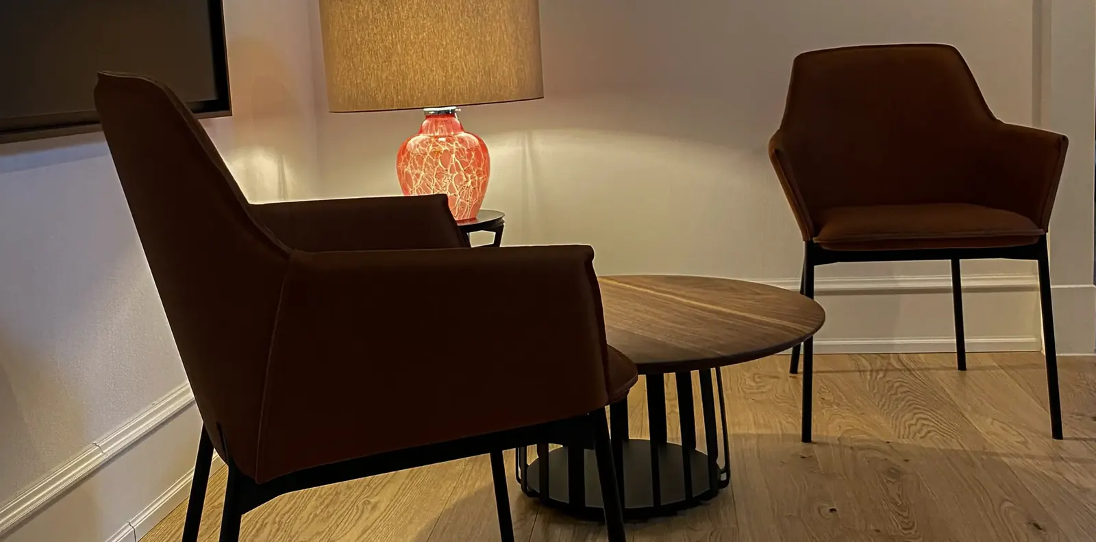 Sitzecke mit zwei braunen Lederarmlehnstühlen um einen runden Holztisch, dahinter eine Leuchte mit orangefarbenem Glasfuß.