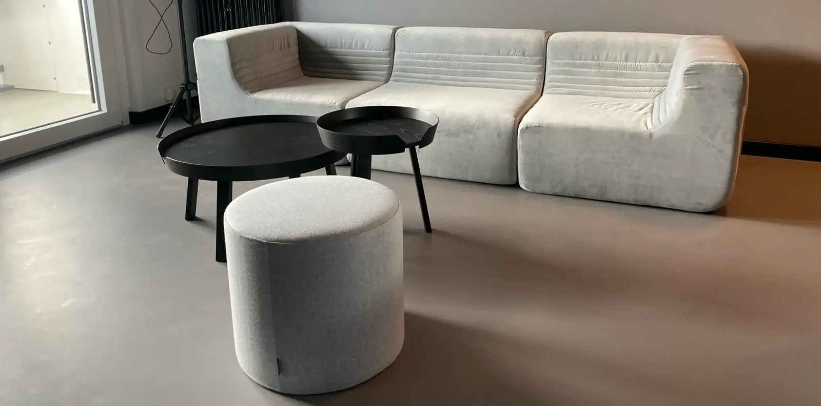 Gemeinschaftsraum mit hellgrauem Modulsofa, zwei runden schwarzen Couchtischen und grauem Sitzhocker auf grauem Boden.