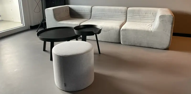 Gemeinschaftsraum mit hellgrauem Modulsofa, zwei runden schwarzen Couchtischen und grauem Sitzhocker auf grauem Boden.