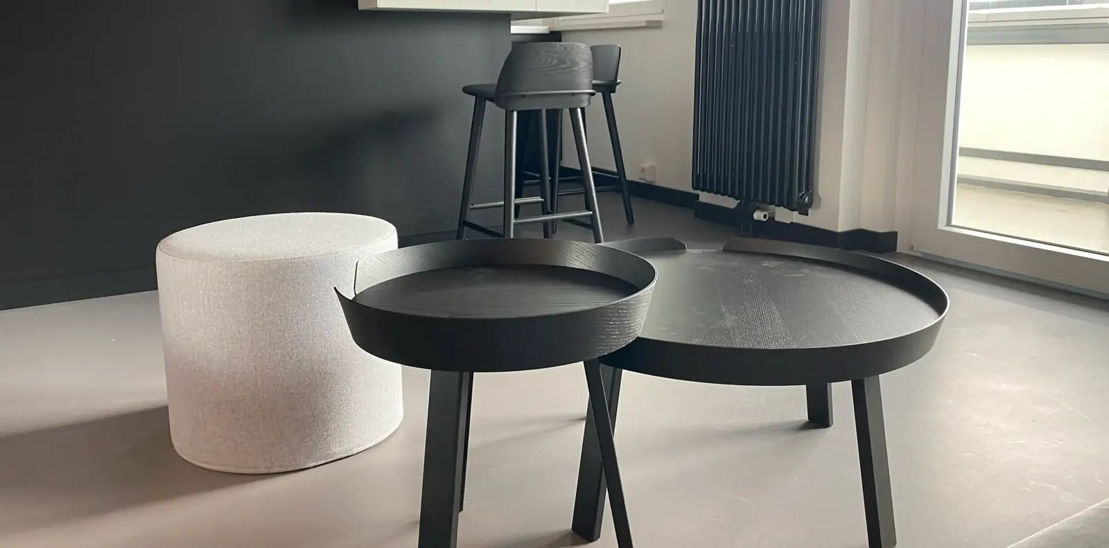 Zwei runde schwarze Holztische mit Tablettrand und ein hellgrauer Polsterhocker vor dunkler Wand, dahinter ein Barhocker.