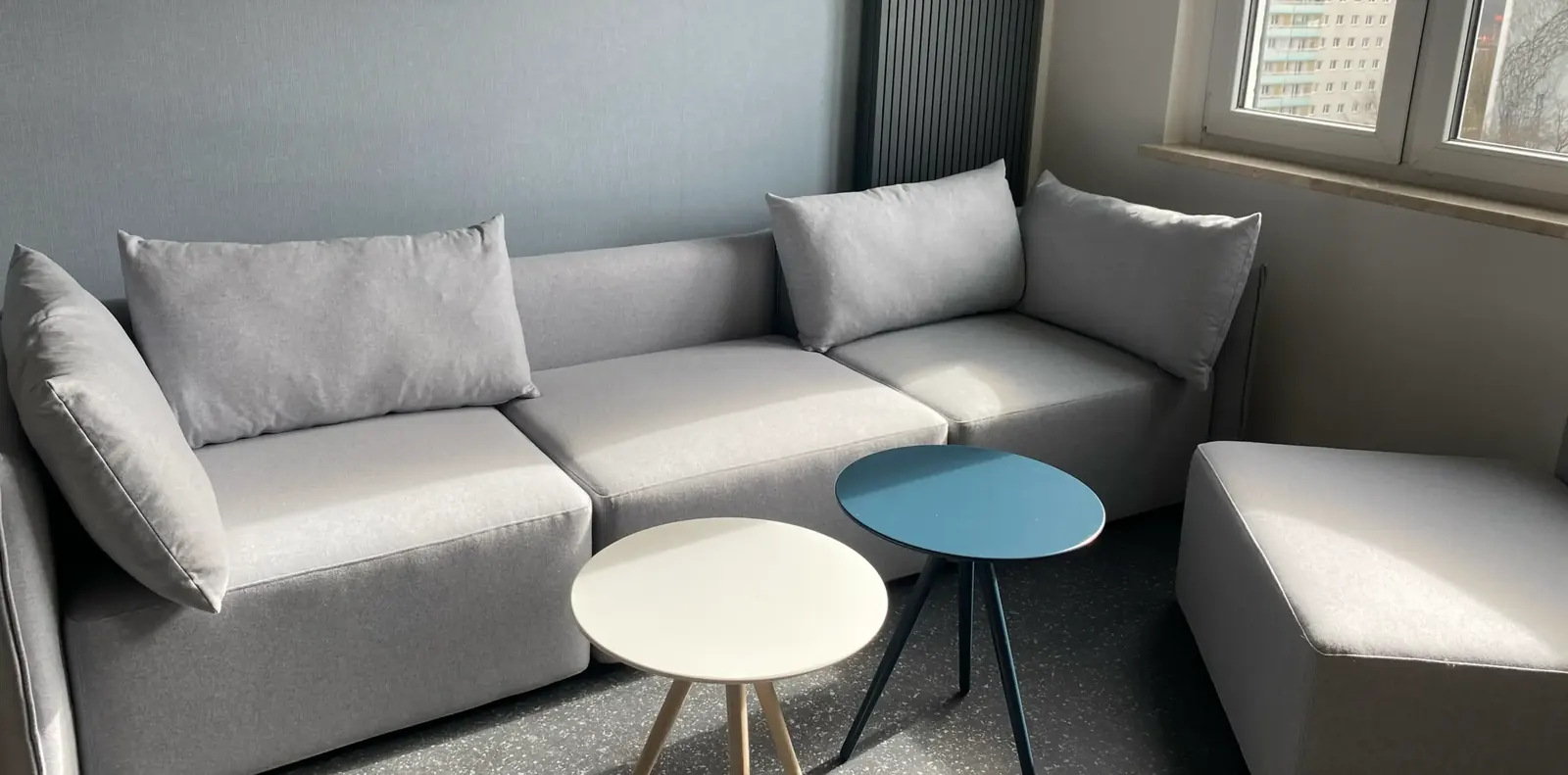 Hellgraues Ecksofa mit Kissen vor blaugrauer Wand, davor zwei kleine runde Beistelltische in Weiß und Petrol.
