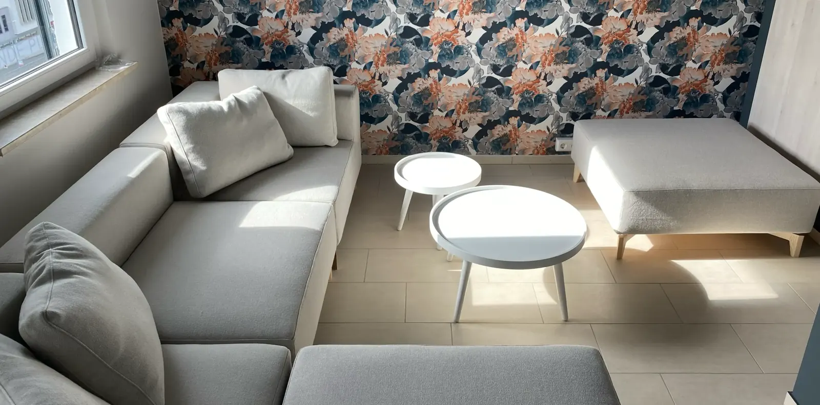 Hellgraues Ecksofa mit Hocker und zwei weißen Beistelltischen vor einer Wand mit großflächiger Blättertapete.