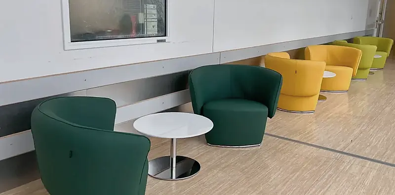 Wartebereich im Flur mit dunkelgrünen und gelben Drehsesseln aus Leder und runden weißen Beistelltischen auf hellem Boden.