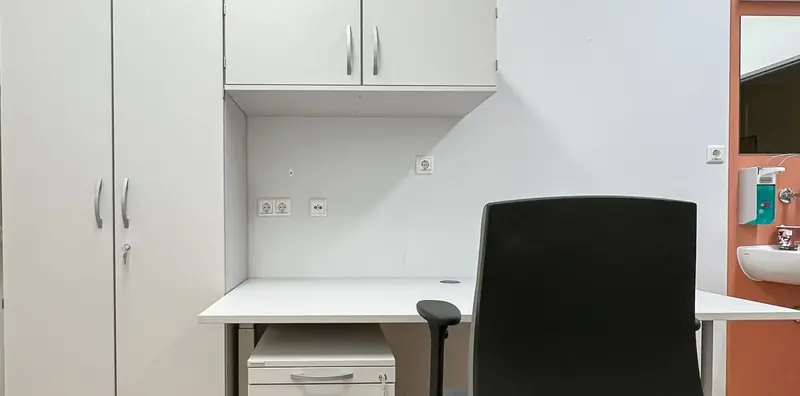 Arbeitsplatz im Behandlungszimmer mit weißem Schreibtisch, Rollcontainer, Hängeschrank und schwarzem Drehstuhl neben einem Waschbecken.