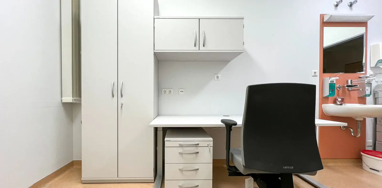 Klinikraum mit weißem Hochschrank, Schreibtisch mit Schubladencontainer, schwarzem Bürodrehstuhl und Waschplatz an oranger Wand.