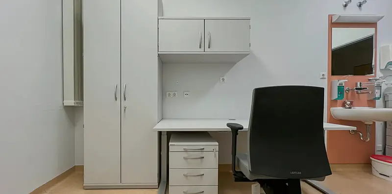 Klinikraum mit weißem Hochschrank, Schreibtisch mit Schubladencontainer, schwarzem Bürodrehstuhl und Waschplatz an oranger Wand.