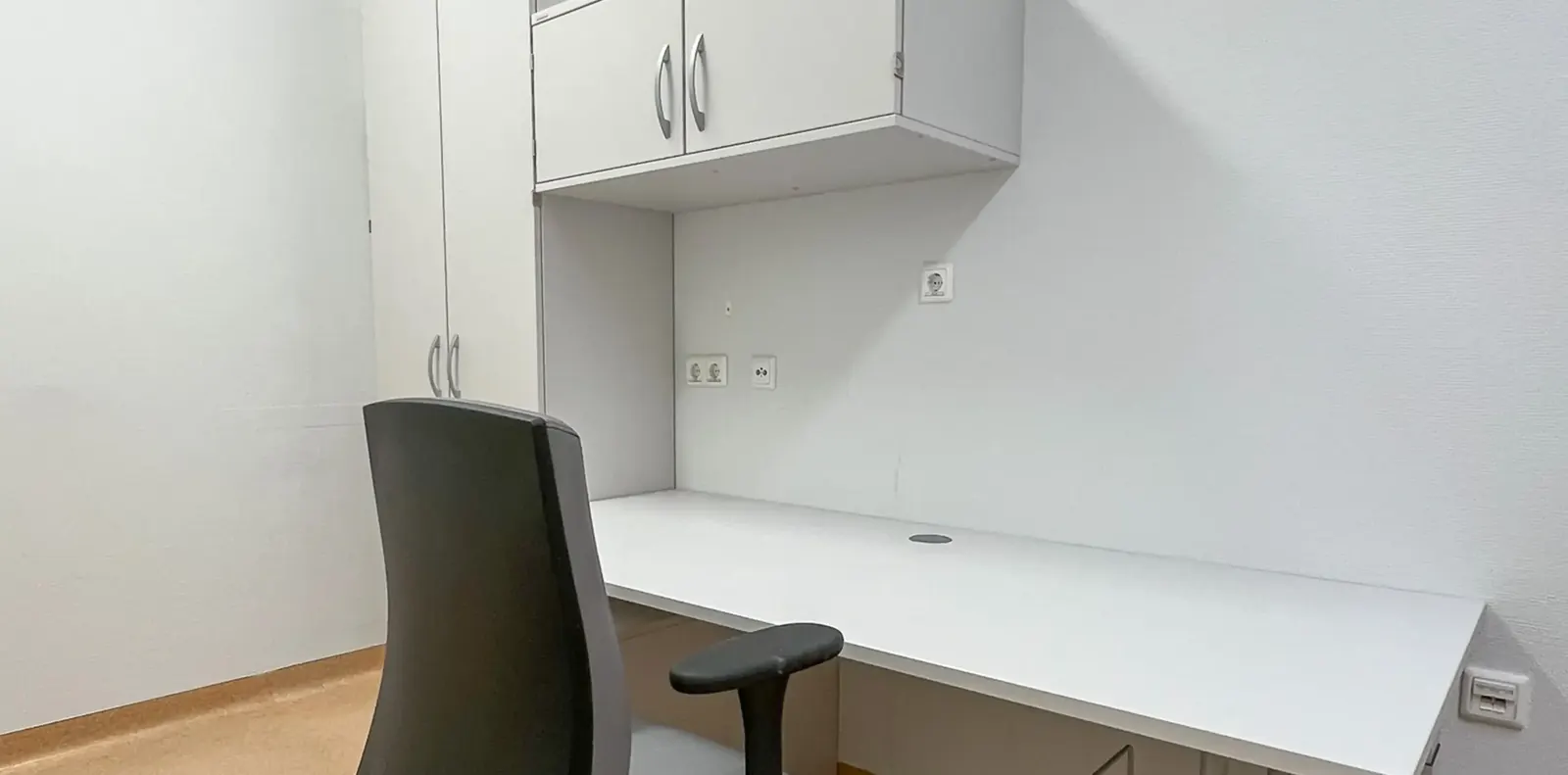 Weißer Schreibtisch über Eck unter Oberschränken mit schwarzem Bürostuhl in einem hellen Klinikzimmer.
