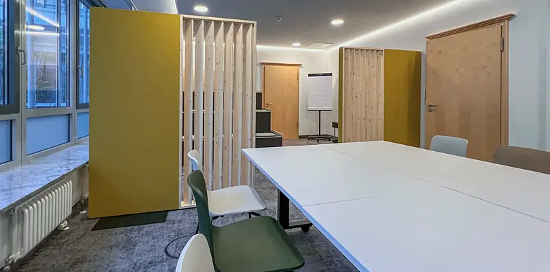 Seminarraum mit langen weißen Tischen, weißen und grünen Schalenstühlen, gelber Akustiktrennwand und Holzlatten-Raumteiler am Fenster.