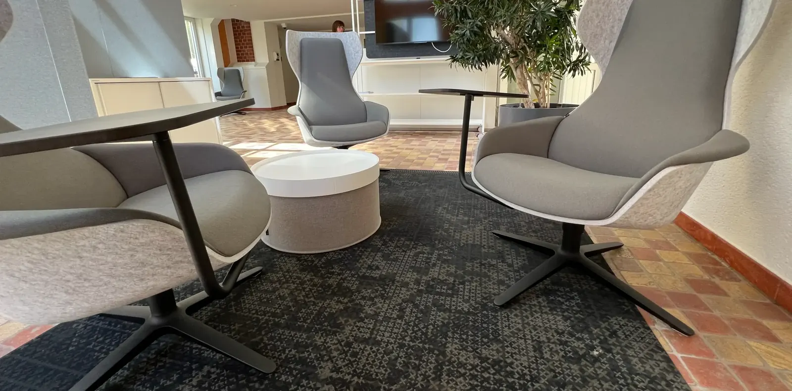 Graue Ohrensessel mit Filzschale und schwenkbarem Ablagetisch um einen weißen runden Hocker auf dunklem Teppich.