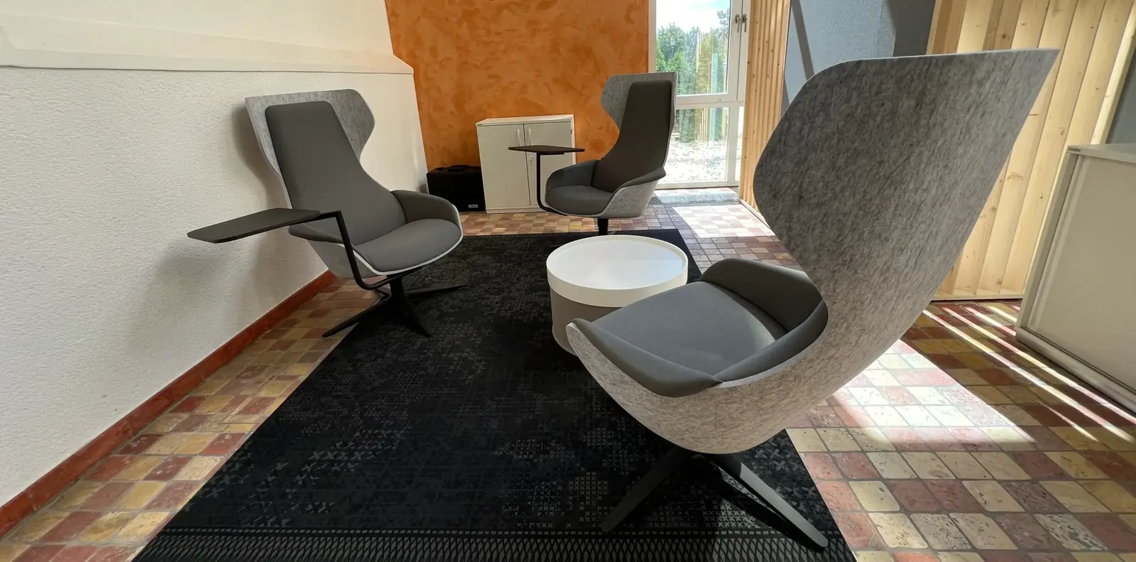 Sitzbereich mit grauen Loungesesseln, Beistelltisch und weißem Rundhocker auf dunklem Teppich über Terrakottafliesen.