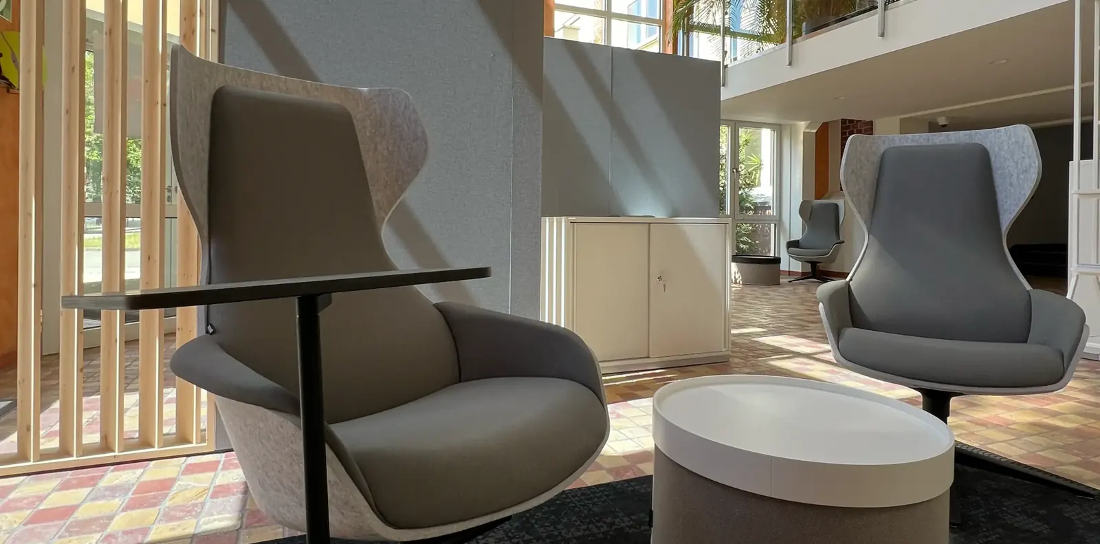 Grauer Loungesessel mit Ablagetisch neben Holzlatten-Raumteiler, dahinter weißer Hocker, zweiter Sessel und Schrank.
