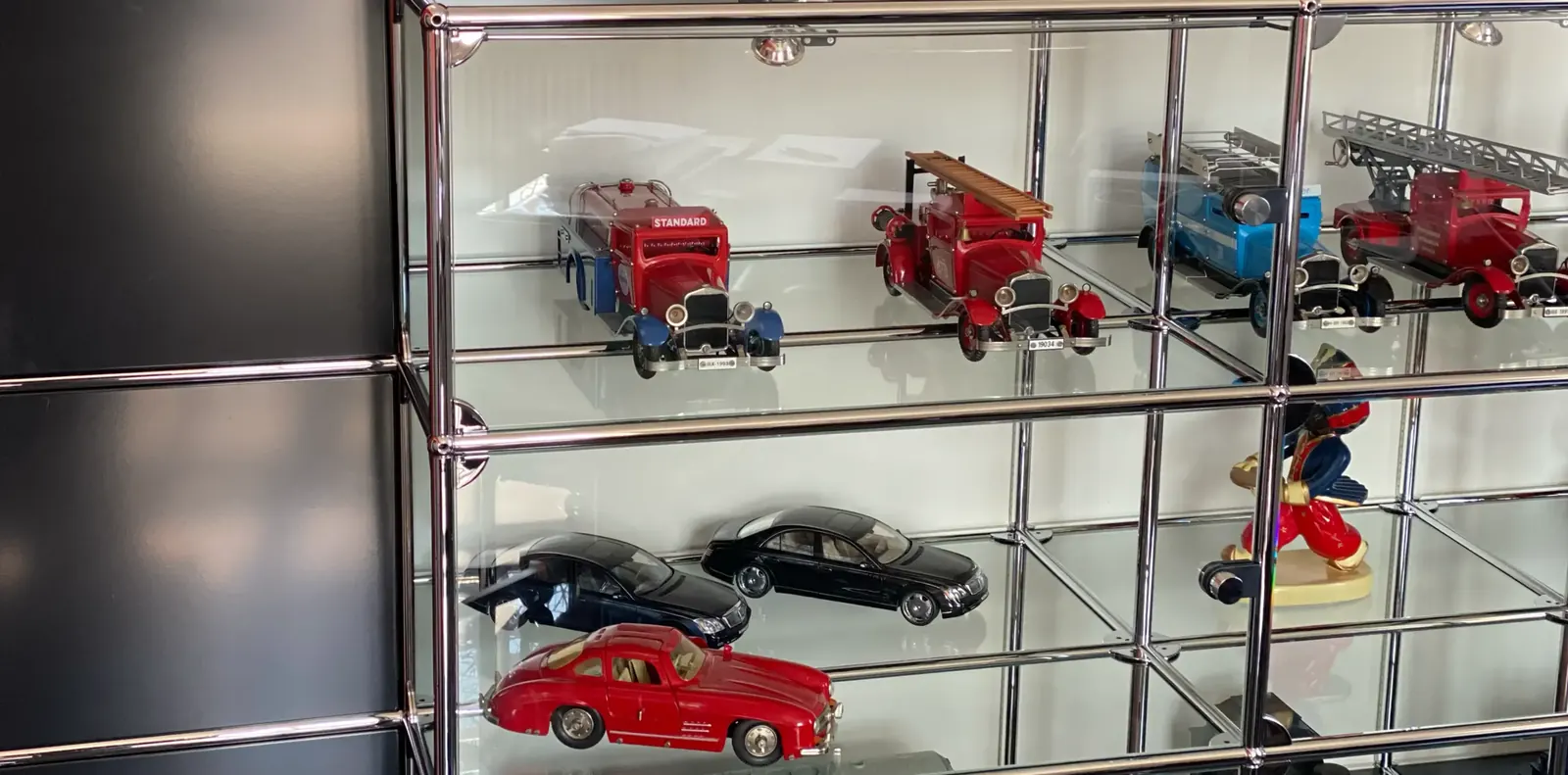 Chromregal mit Glasböden als Vitrine für Modellautos und Feuerwehrfahrzeuge neben schwarzen Metallfronten.