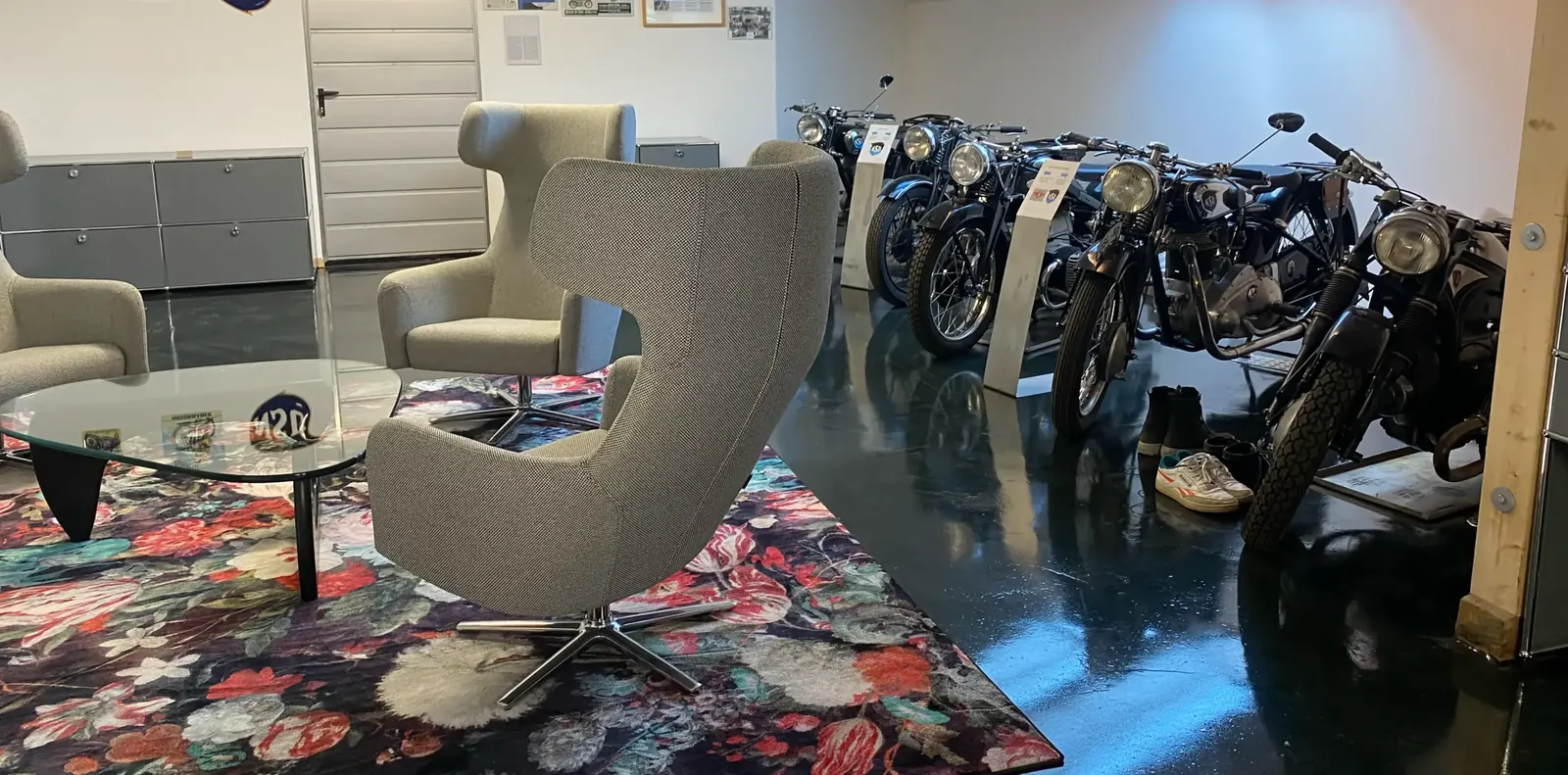 Graue Drehsessel und Glastisch auf Blumenteppich neben einer Reihe ausgestellter Oldtimer-Motorräder auf dunklem Boden.