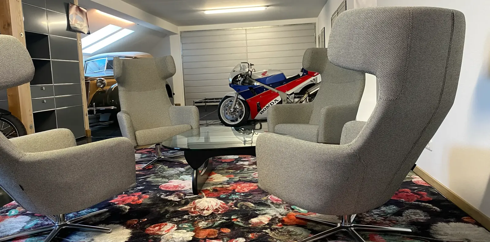 Vier graue Ohrensessel um einen Glascouchtisch, im Hintergrund ein Rennmotorrad, ein Oldtimer und graue Regale.