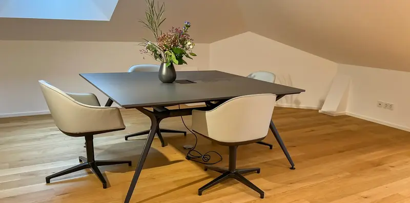 Anthrazitfarbener Besprechungstisch mit Blumenvase und vier cremefarbenen Drehsesseln auf Sternfuß unter Dachschräge.