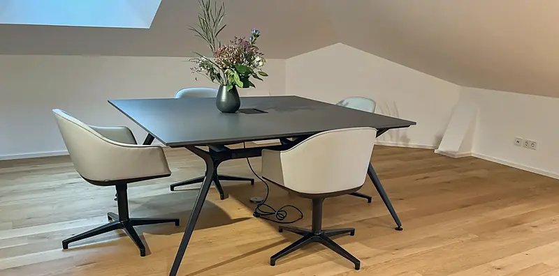 Anthrazitfarbener Besprechungstisch mit Blumenvase und vier cremefarbenen Drehsesseln auf Sternfuß unter Dachschräge.