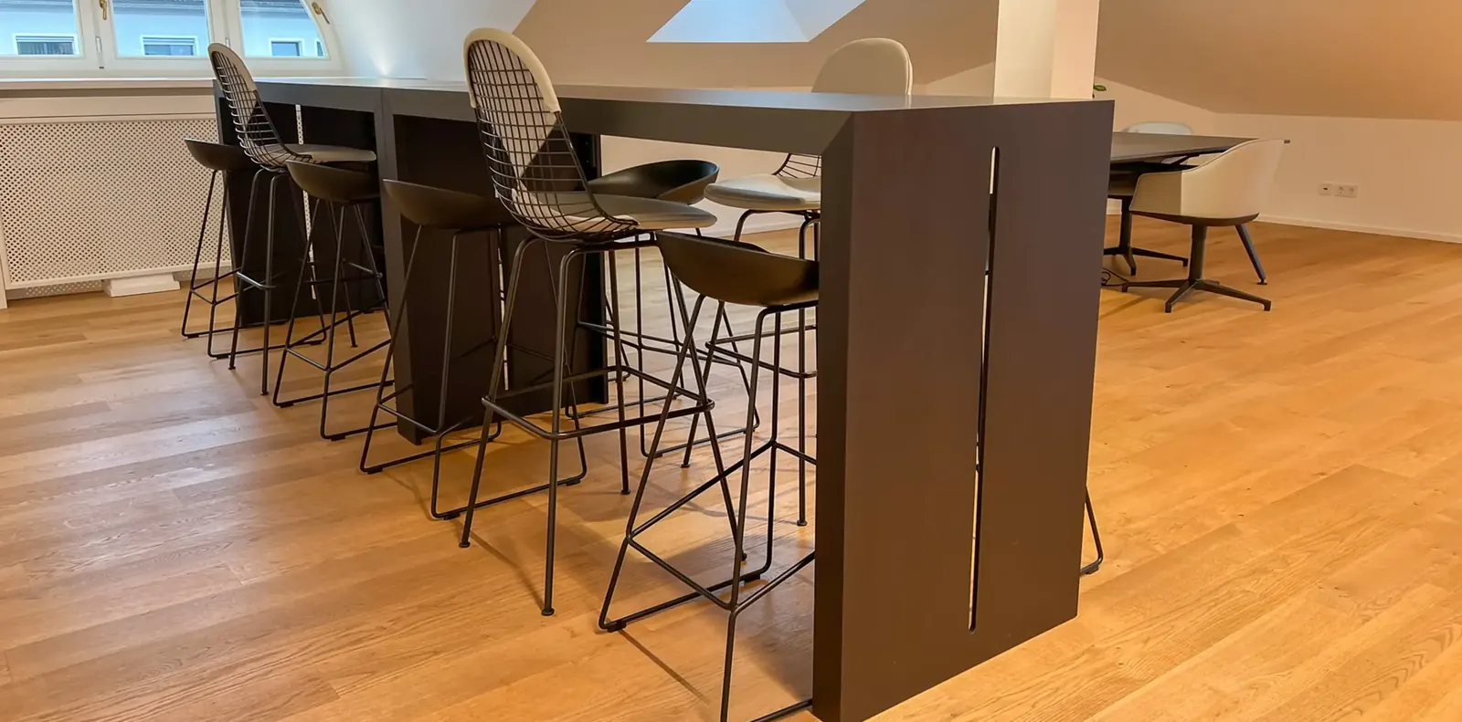 Dunkler Stehtisch mit Wangen aus Holz und Barhockern mit Drahtgestell und schwarzen Sitzschalen auf Eichenparkett.