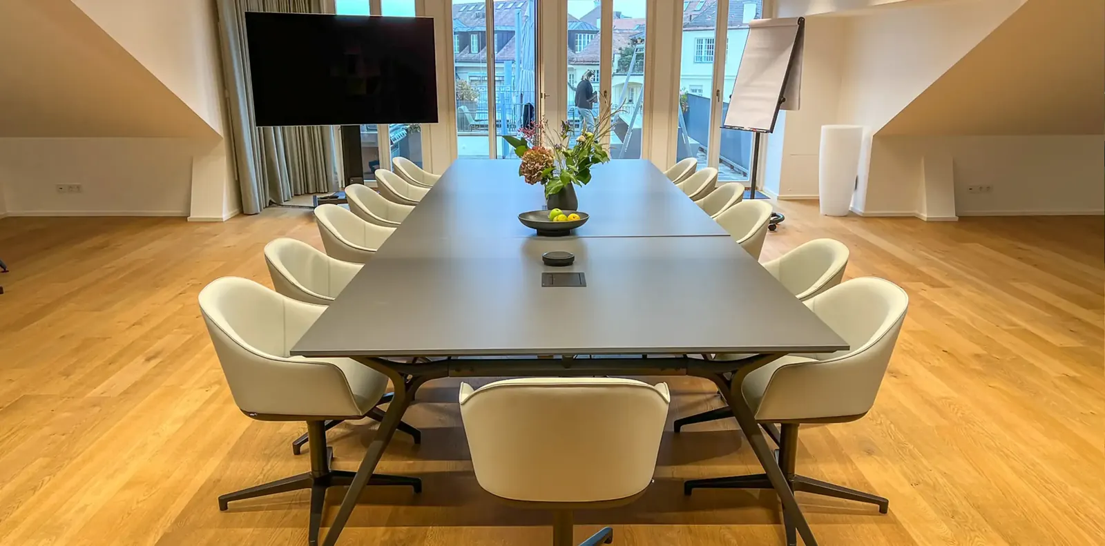 Frontalansicht einer langen Konferenztafel mit zwölf cremefarbenen Schalensesseln, hinten Fenstertüren und Flipchart.