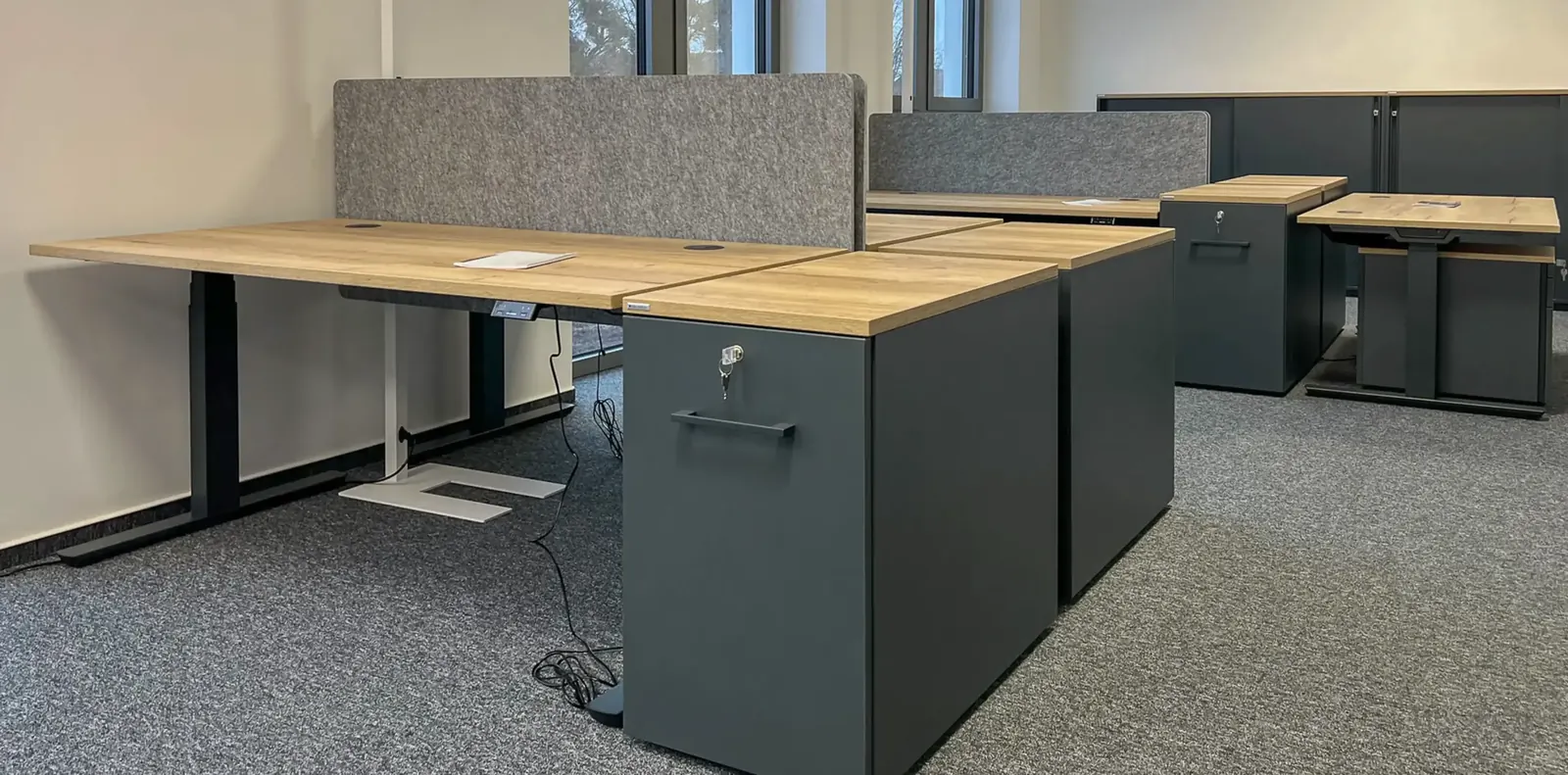 Höhenverstellbare Schreibtische in Eichenoptik mit Filz-Sichtschutz und abschließbaren anthrazitfarbenen Containern.