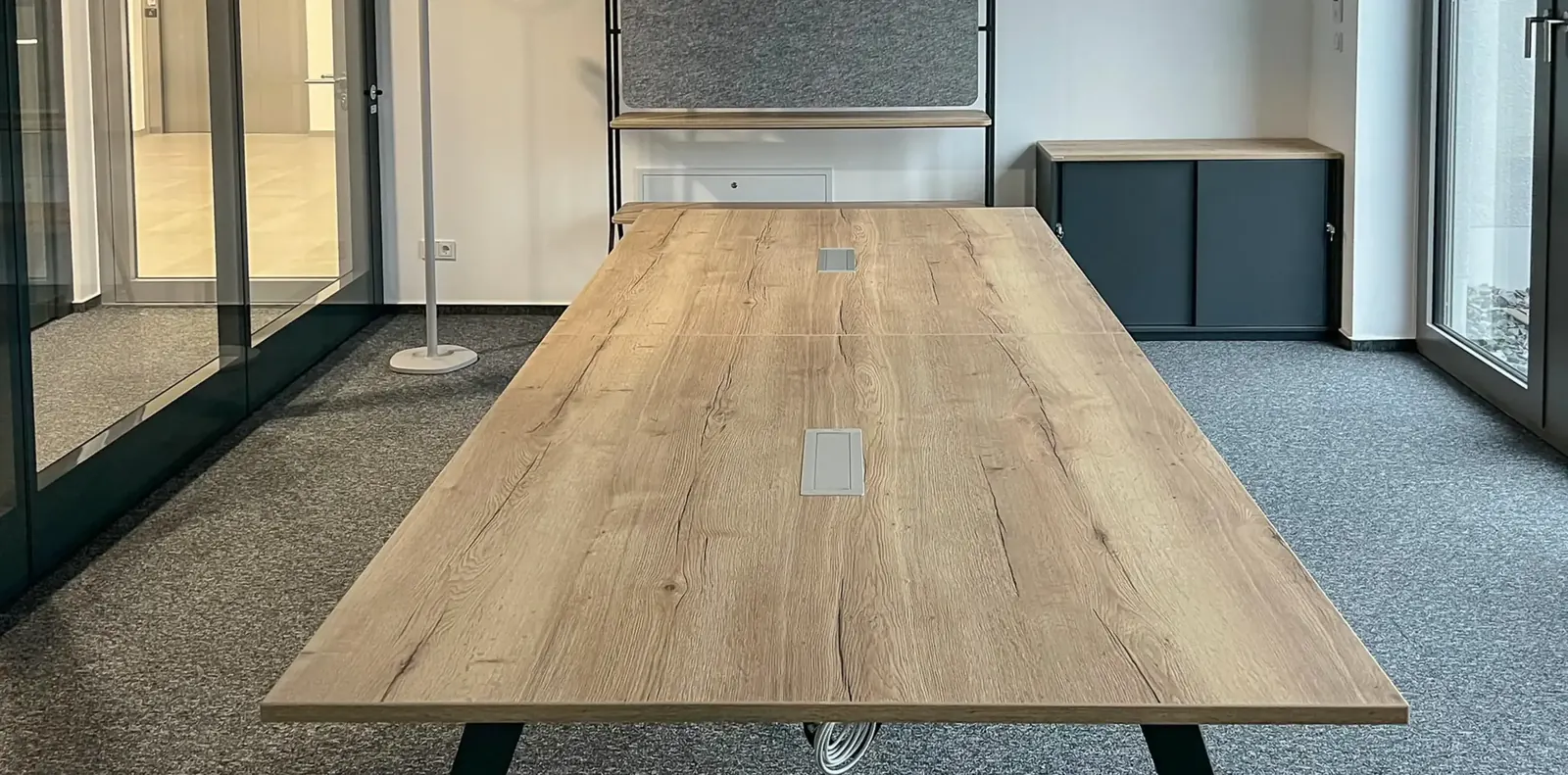 Langer Besprechungstisch in Eichenoptik mit Kabeldurchlässen auf grauem Teppich, dahinter ein anthrazitfarbener Schrank.