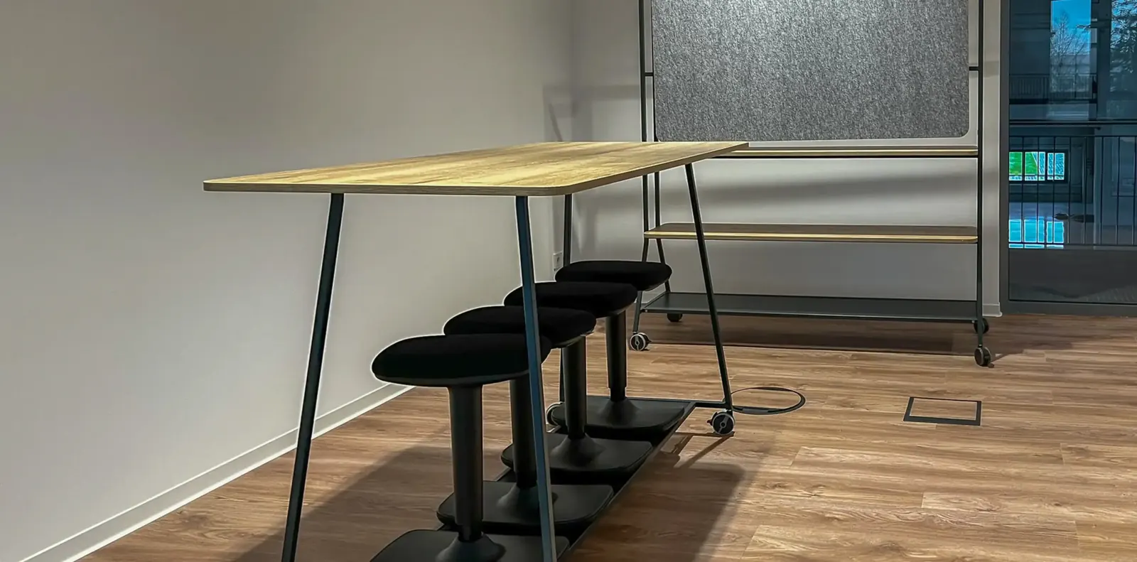 Stehtisch in Holzoptik mit schwarzen Wipphockern auf Holzboden, dahinter ein Regal mit Filzrückwand.