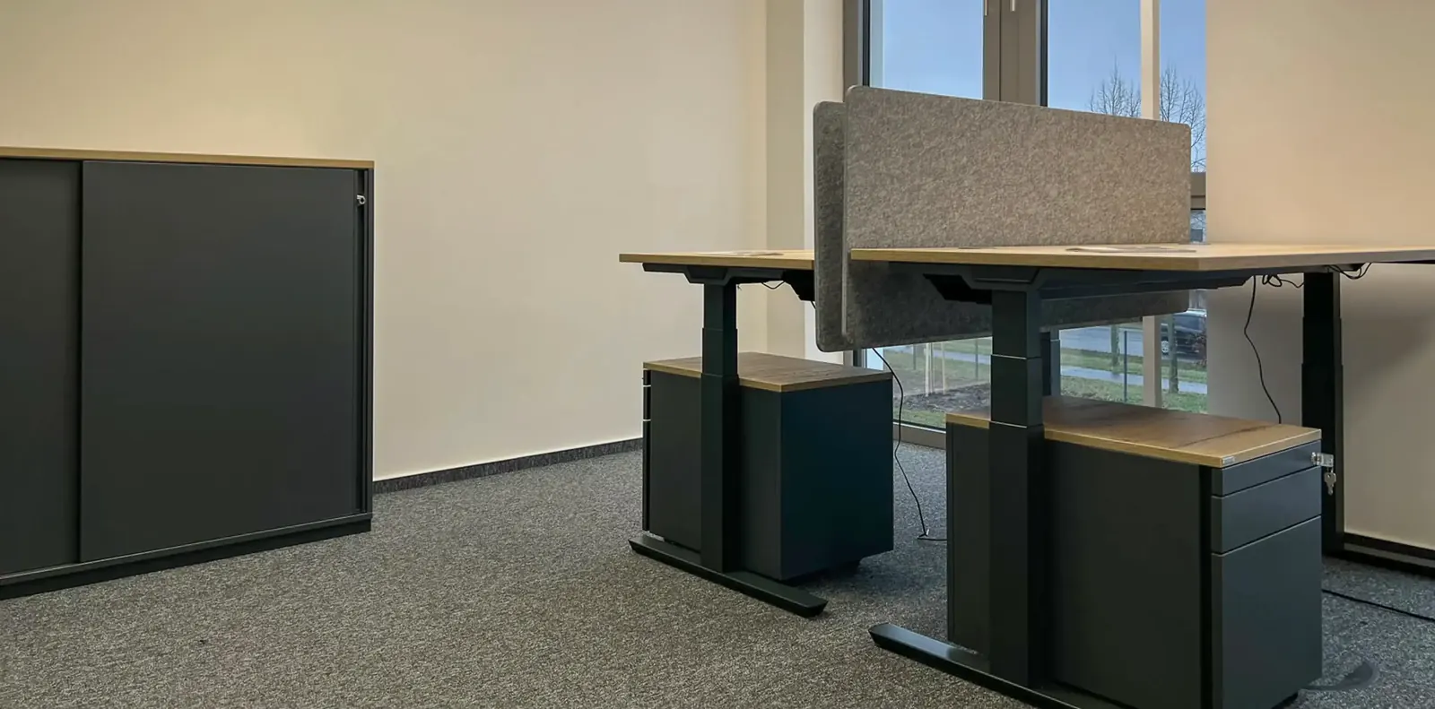 Höhenverstellbare Schreibtische mit Filz-Sichtschutz und Rollcontainern neben einem anthrazitfarbenen Schiebetürenschrank.
