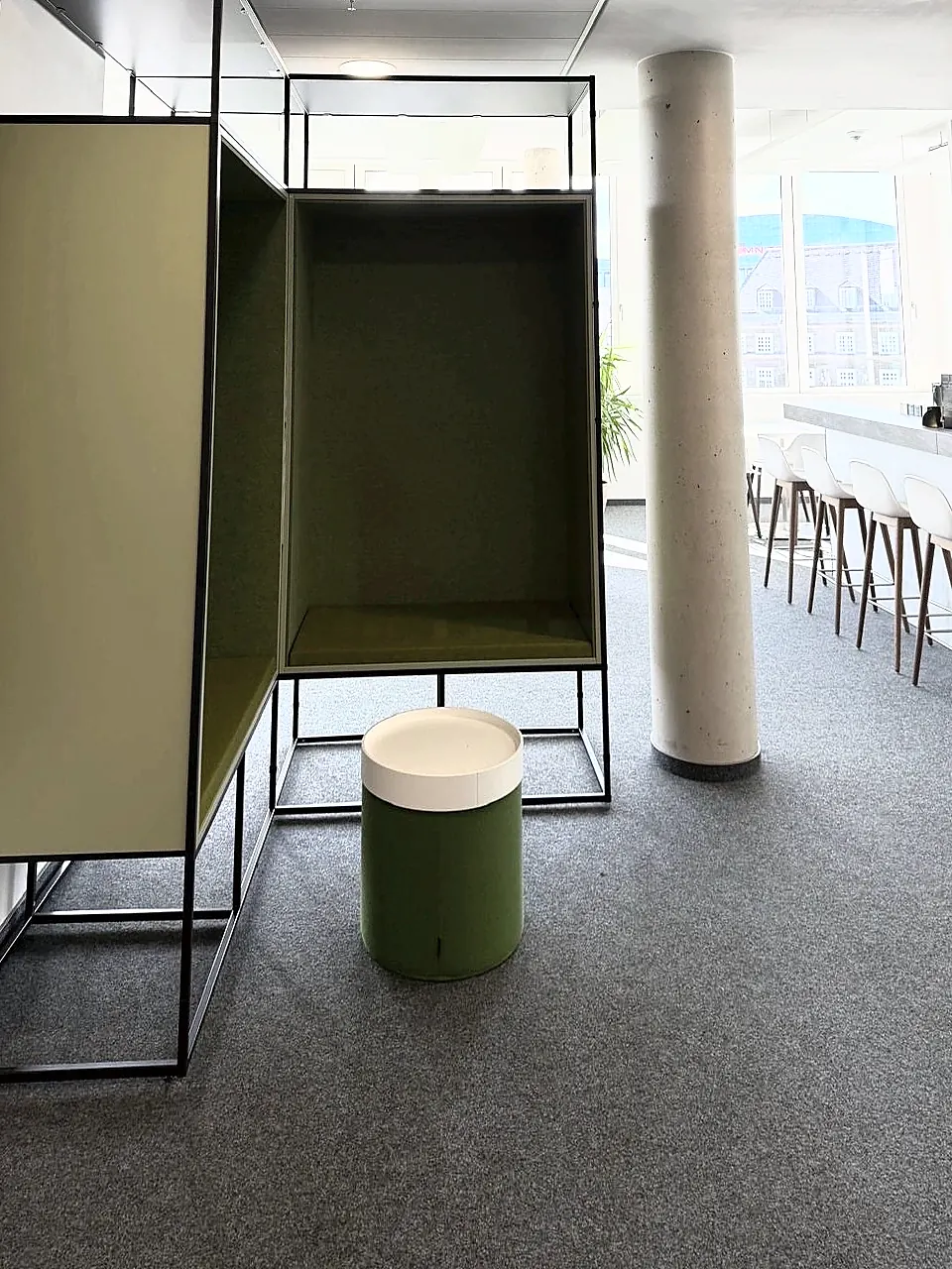 Eckige Sitznische mit olivgrünen Polsterwänden in schwarzem Rahmen, davor ein grüner Hocker mit heller Sitzfläche, im Hintergrund eine Bartheke.