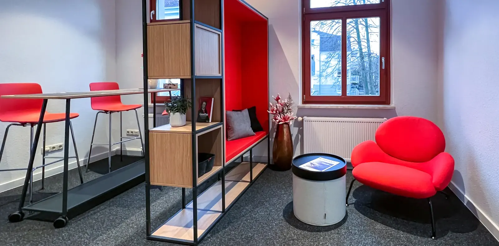 Raum mit roten Barhockern an einem Stehtisch, offenem Regal als Raumteiler, roter Sitzbank und rotem Loungesessel.