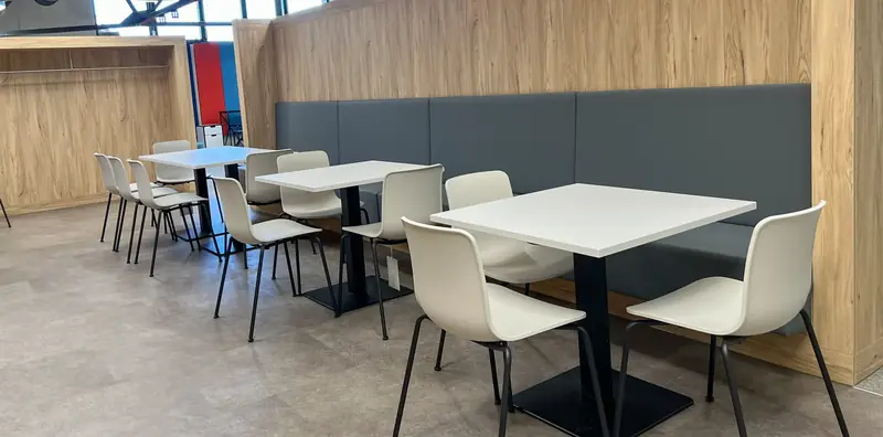 Aufenthaltsraum mit Holzwandverkleidung, grauer Polsterbank über Eck, weißen Tischen und cremefarbenen Schalenstühlen.