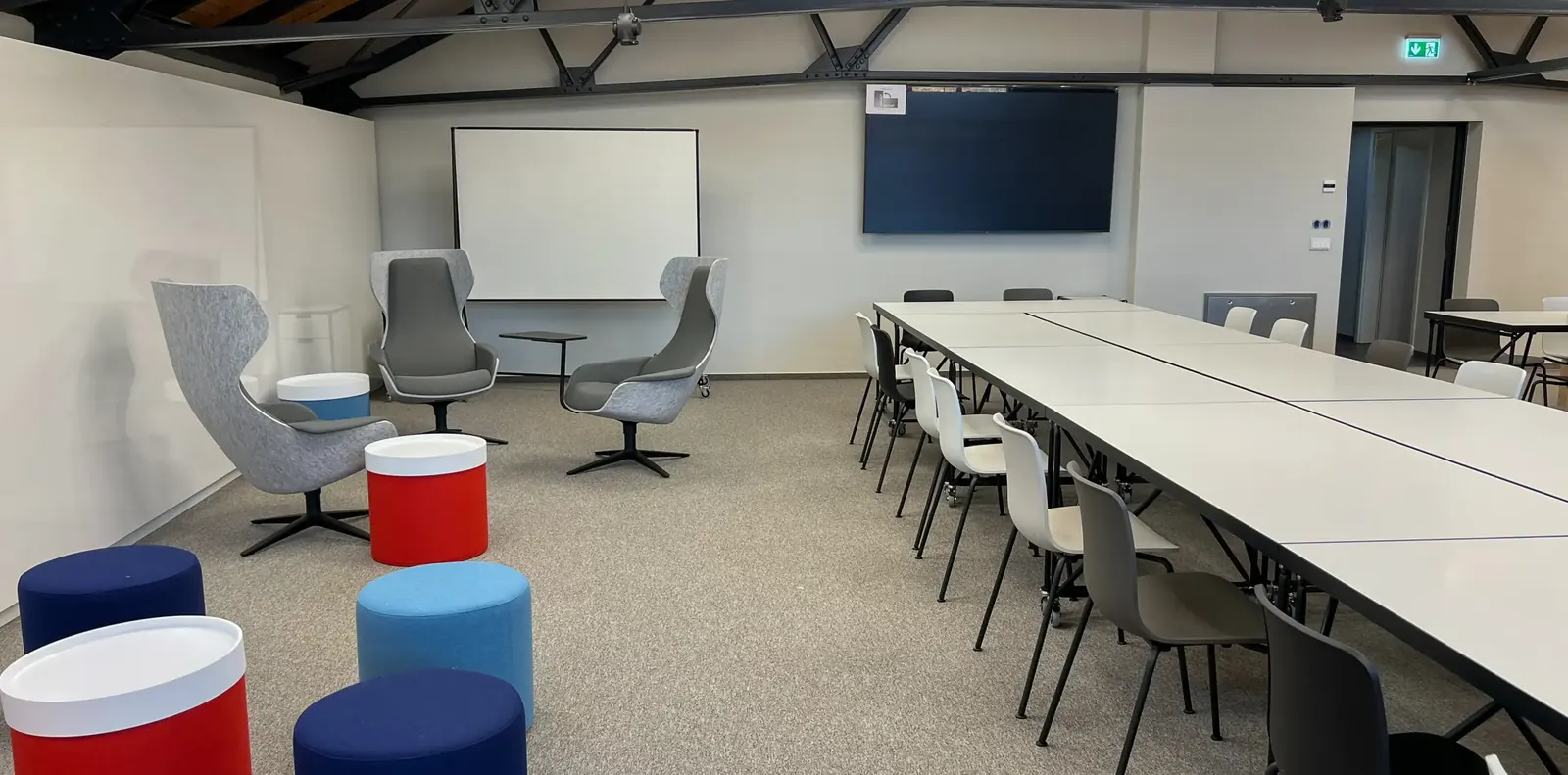 Schulungsraum mit langer Tischreihe und Schalenstühlen, vorne bunte Sitzhocker, hinten graue Ohrensessel und Whiteboard.