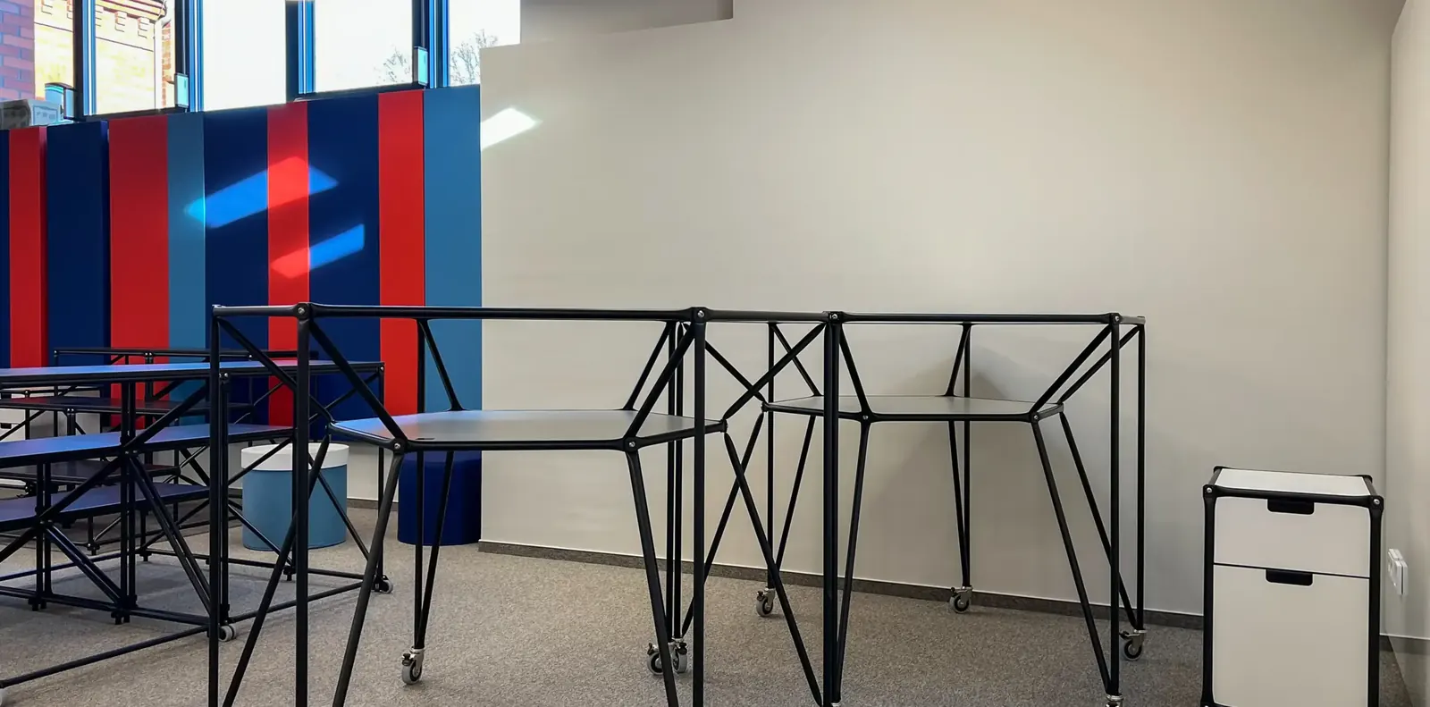 Schwarze Stehtische aus Stahlrohr auf Rollen vor rot-blau gestreiften Akustikpaneelen, rechts ein weißer Rollcontainer.