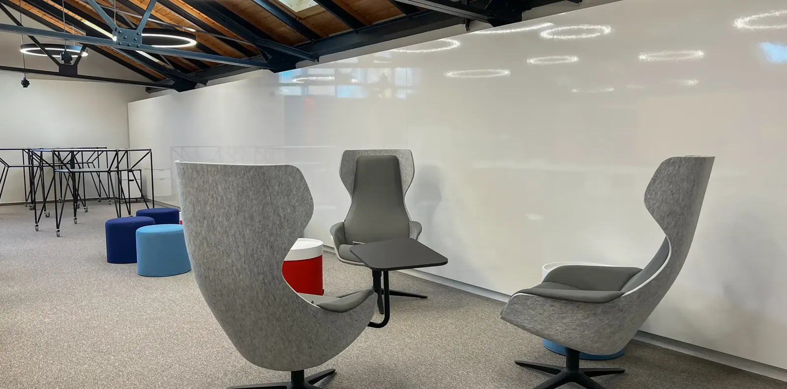 Drei graue Ohrensessel mit Sternfuß und Ablagetisch vor einer Whiteboardwand, dahinter blaue Sitzhocker und Stahlgestelle.
