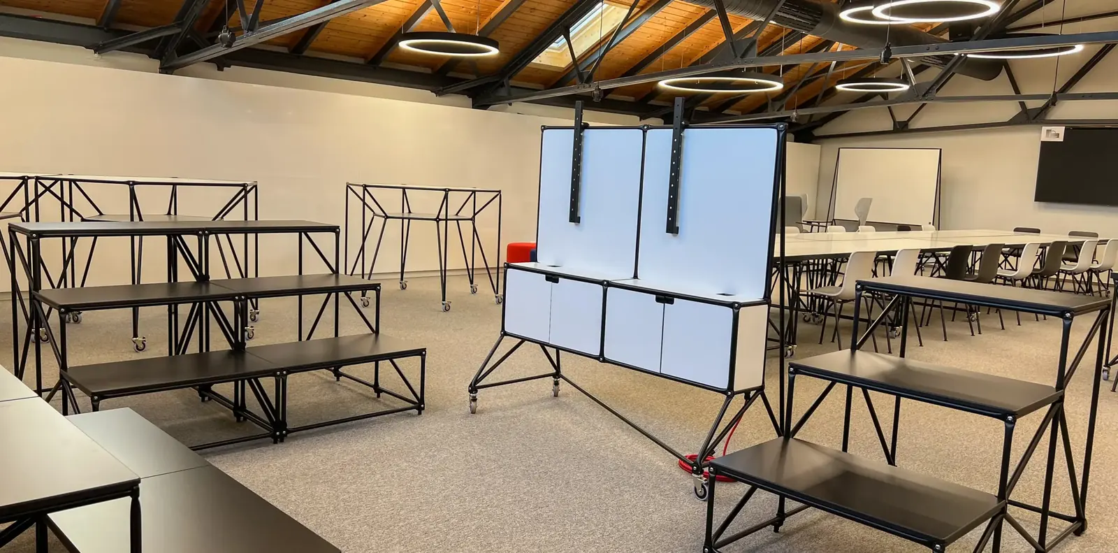 Fahrbare Whiteboard-Stellwand und schwarze System-180-Sitzpodeste in einer Halle mit Ringleuchten und Tischreihen.