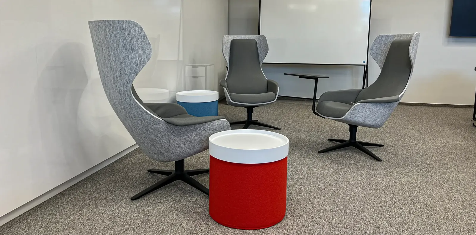 Loungeecke mit drei grauen Ohrensesseln auf Sternfuß, rotem Hocker mit weißem Deckel und Whiteboard an der Wand.