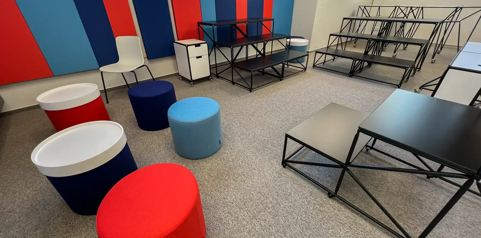 Runde Filzhocker in Rot, Blau und Hellblau vor Wandpaneelen in Rot und Blau, rechts schwarze Stahlrohr-Sitzstufen.