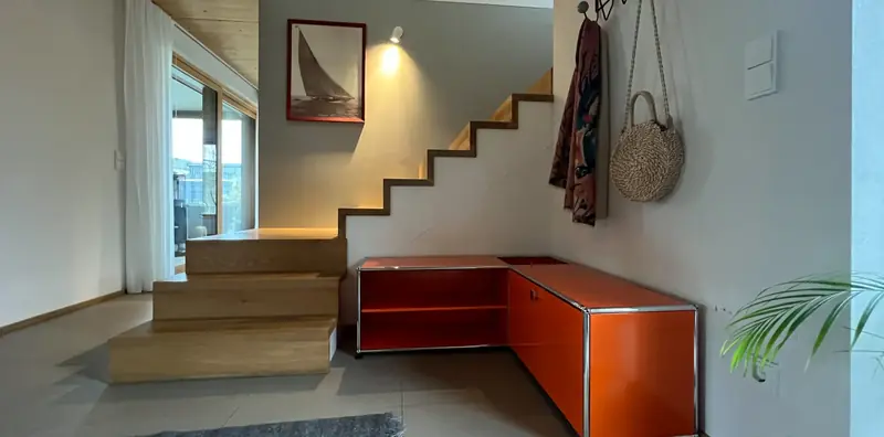 Flur mit Holztreppe und orangefarbenem USM-Sideboard über Eck, darüber ein Vitra-Garderobenhaken mit Tasche und Tuch.