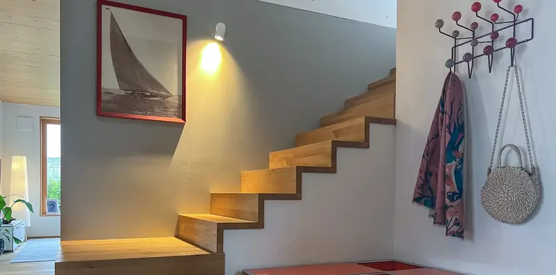 Beleuchtete Eichentreppe an grauer Wand mit Segelbootbild, rechts eine Wandgarderobe mit bunten Kugelhaken.