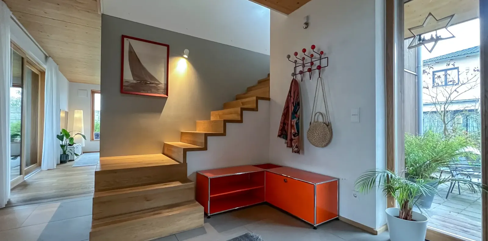 Eingangsbereich mit Treppe, orangefarbenem USM-Möbel über Eck und Wandgarderobe, rechts Blick durch die Glastür auf die Terrasse.