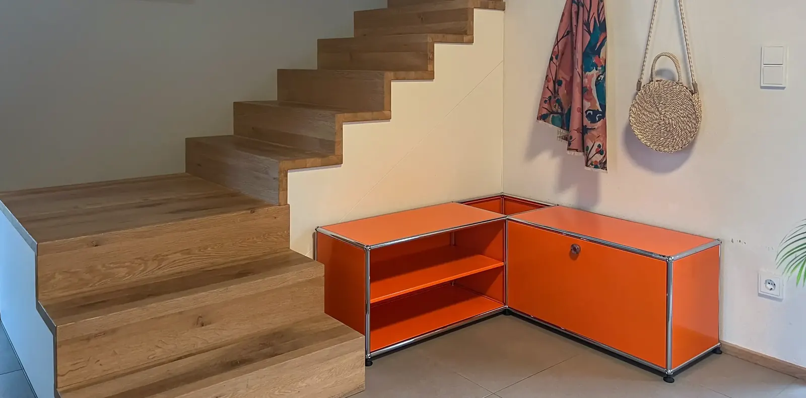 Orangefarbenes USM-Eckmöbel mit offenem Regalfach und Klappe neben den unteren Stufen einer Eichentreppe.
