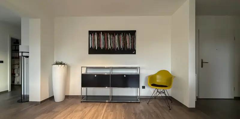 Wohnraum mit schwarzem USM-Sideboard mit Chromgestell, gelbem Vitra-Schalenstuhl und weißem Pflanzkübel vor heller Wand.