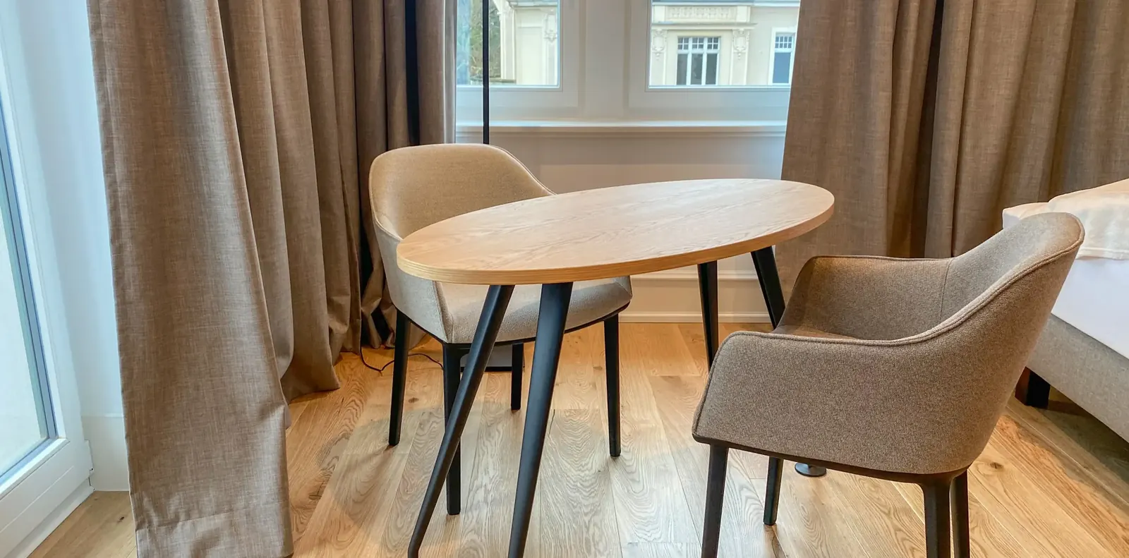 Ovaler Holztisch mit schwarzen Beinen und zwei beigen Vitra-Polsterstühlen am Fenster mit bodenlangen Leinenvorhängen.