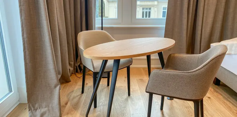 Ovaler Holztisch mit schwarzen Beinen und zwei beigen Vitra-Polsterstühlen am Fenster mit bodenlangen Leinenvorhängen.