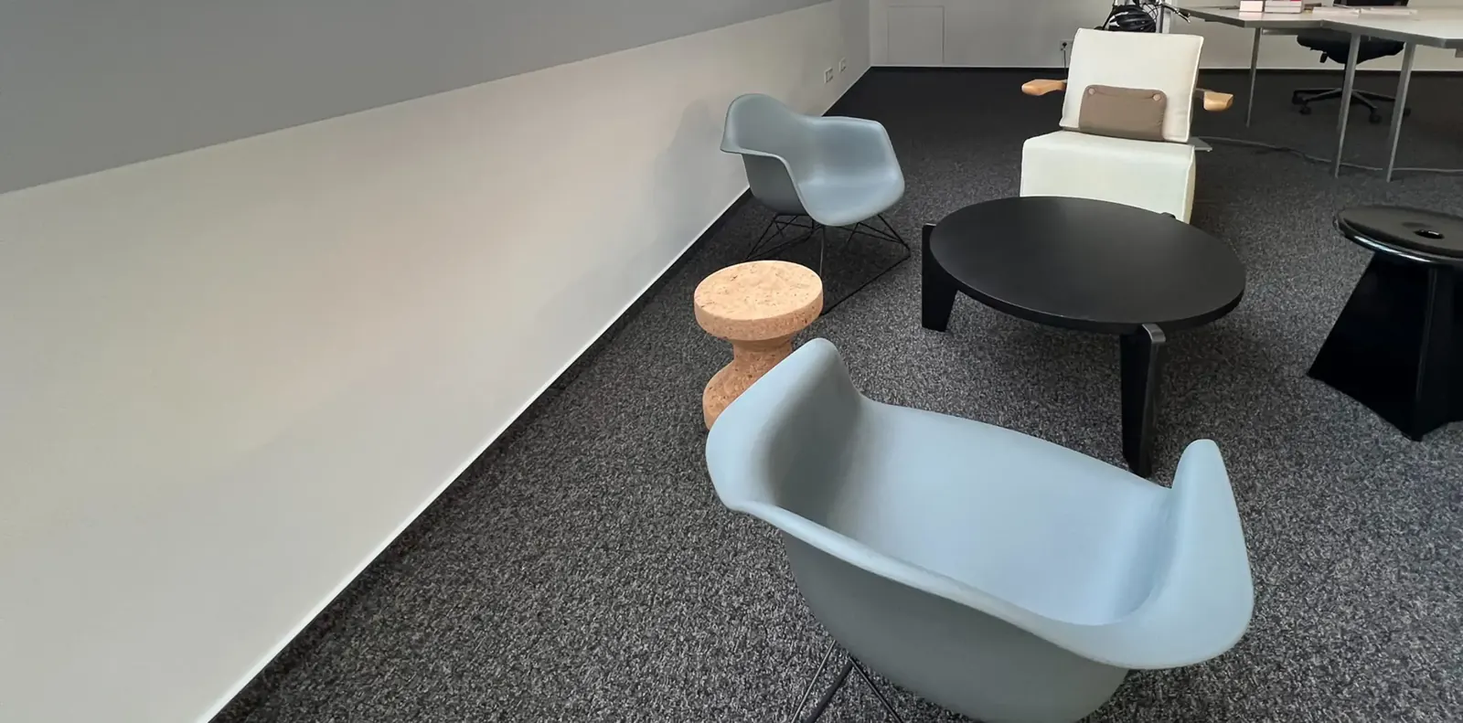 Loungebereich auf dunklem Teppich mit hellblauen Vitra-Schalensesseln, Korkhocker, rundem schwarzem Couchtisch und weißem Sessel.