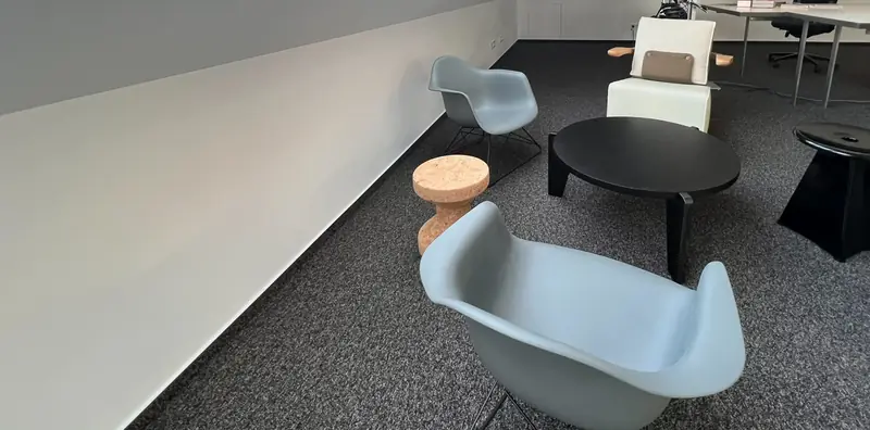 Loungebereich auf dunklem Teppich mit hellblauen Vitra-Schalensesseln, Korkhocker, rundem schwarzem Couchtisch und weißem Sessel.
